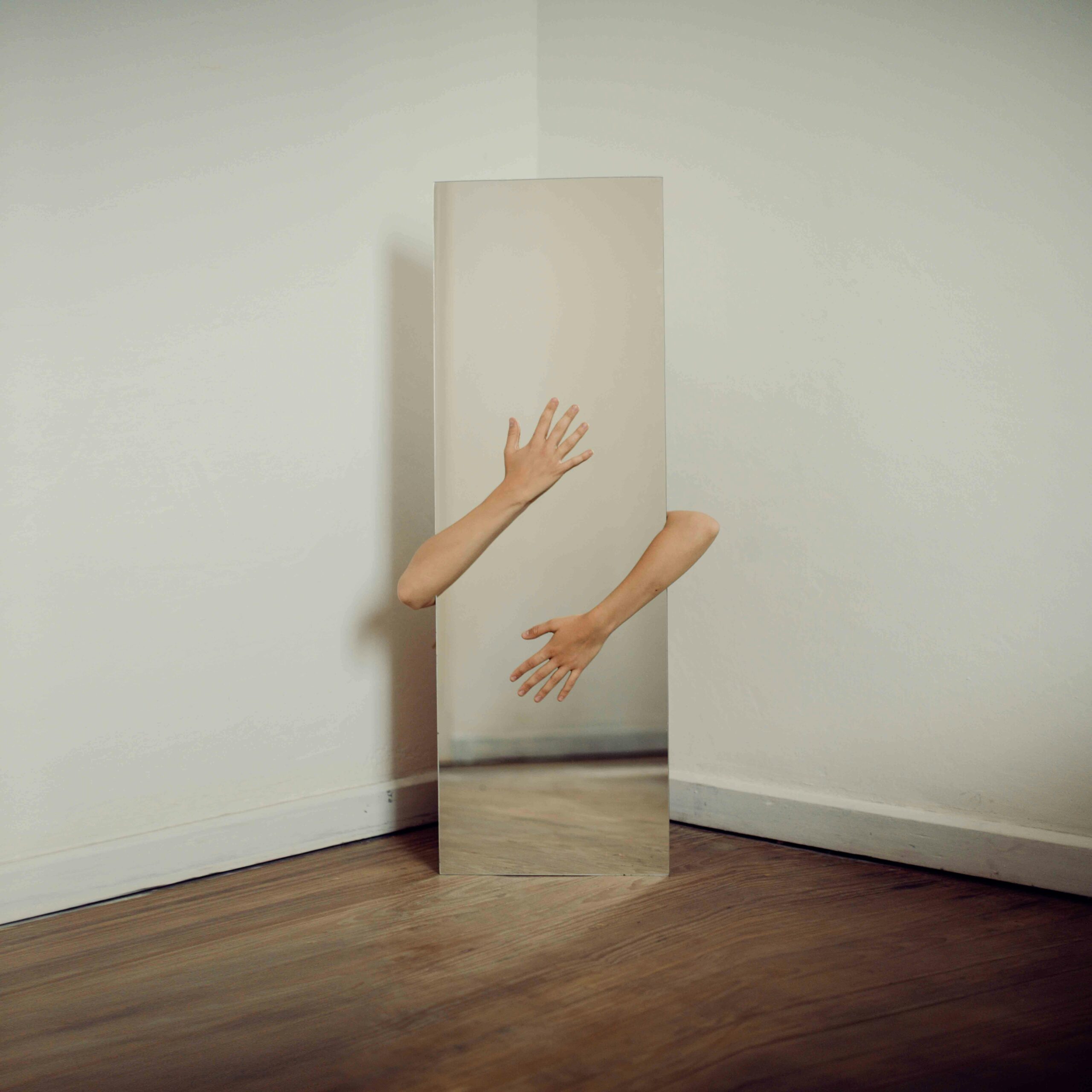 Two arms reach around a mirror set in the corner of a blank white room
