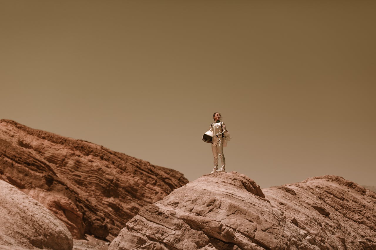 A lone astronaut stands on a barren rock face, the sky brown behind her, holding her helmet against her hip