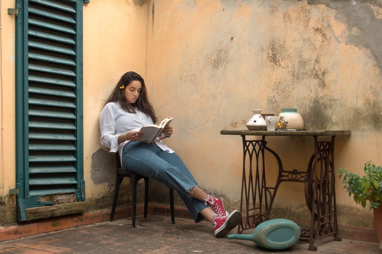 A girl with curly dark brown hair leans against the wall on a stool reading. The wall is peach stucco with a green french blind next to her. She wears a white collared shirt and blue jeans.