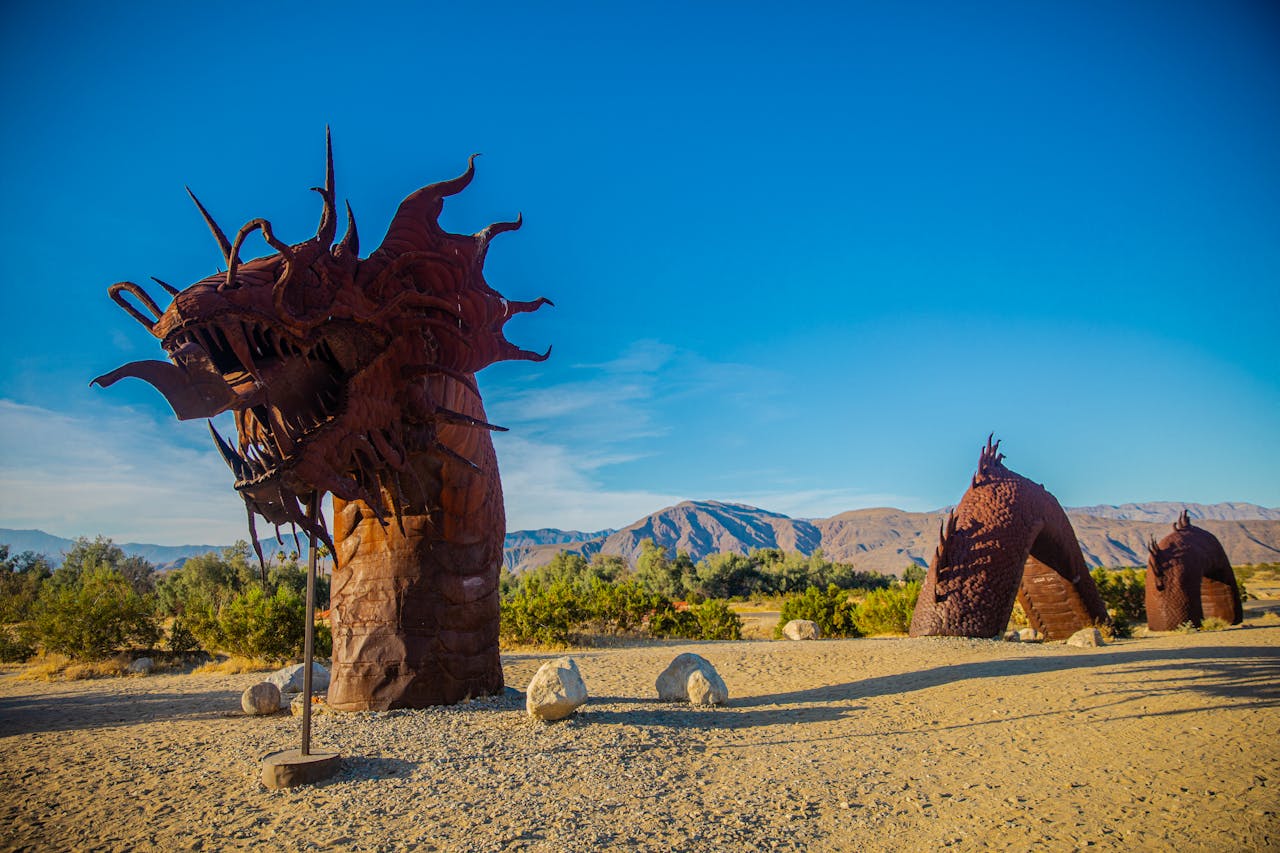A brown metal Asian dragon sculpture; its head sticks out of sand in a desert. Two coils raise up from the ground further back. Mountains and a blue sky in the background.