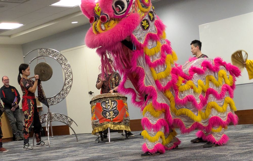 A pink lion dancer stretches its neck up in the foreground; behind, a barrel-sized drum and a gong on a crescent holder and some cymbal players with scarves off the ends of the cymbals
